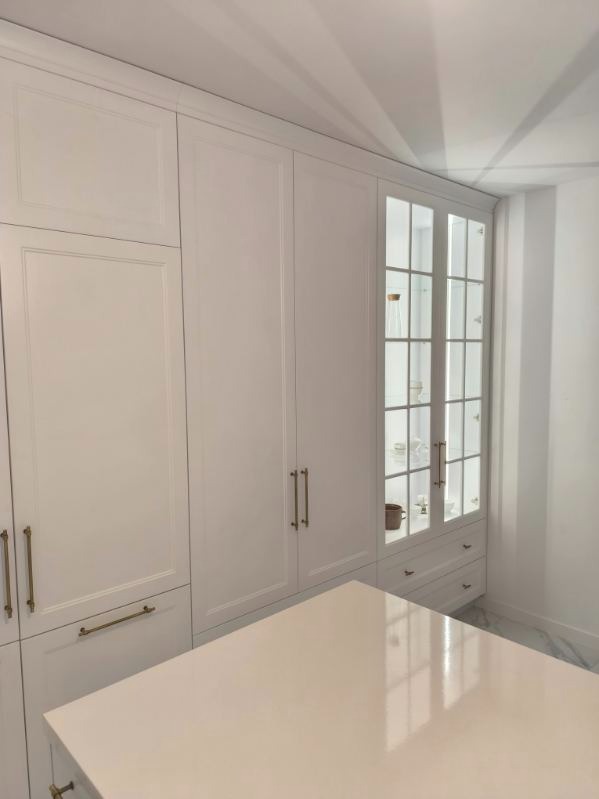 Biała zabudowa kuchenna z przeszkloną witryną i złotymi uchwytami, uzupełniona o blat. Minimalistyczny design, subtelne oświetlenie, marmurowa podłoga.