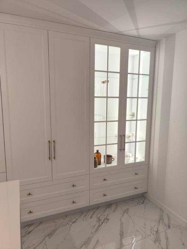 Biała zabudowa meblowa z przeszkloną witryną, podświetloną od wewnątrz. Widoczne naczynia i dekoracje. Marmurowa podłoga. Ujęcie z dołu.