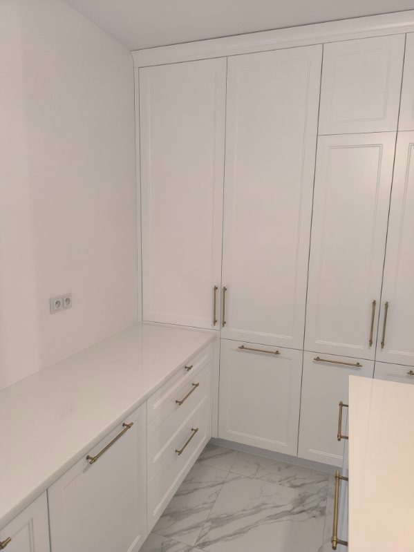 Biała zabudowa kuchenna z frezowanymi frontami i złotymi uchwytami, uzupełniona blatem z konglomeratu, na podłodze marmurowe płytki. Minimalistyczny design.