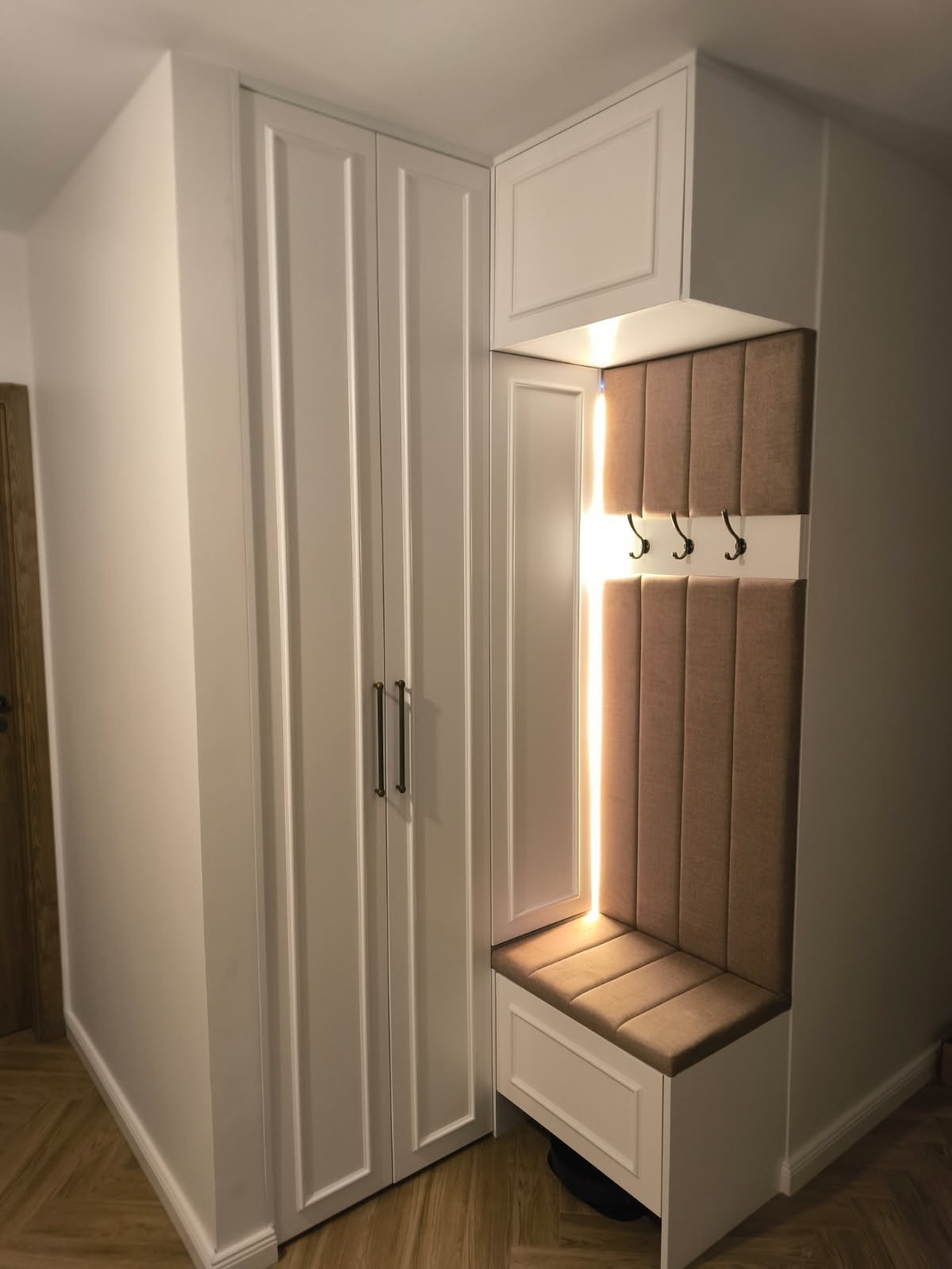 Biała szafa wnękowa z siedziskiem i wieszakami w przedpokoju, z delikatnym oświetleniem i tapicerowanym panelem na ścianie, widok z boku, drewniana podłoga.