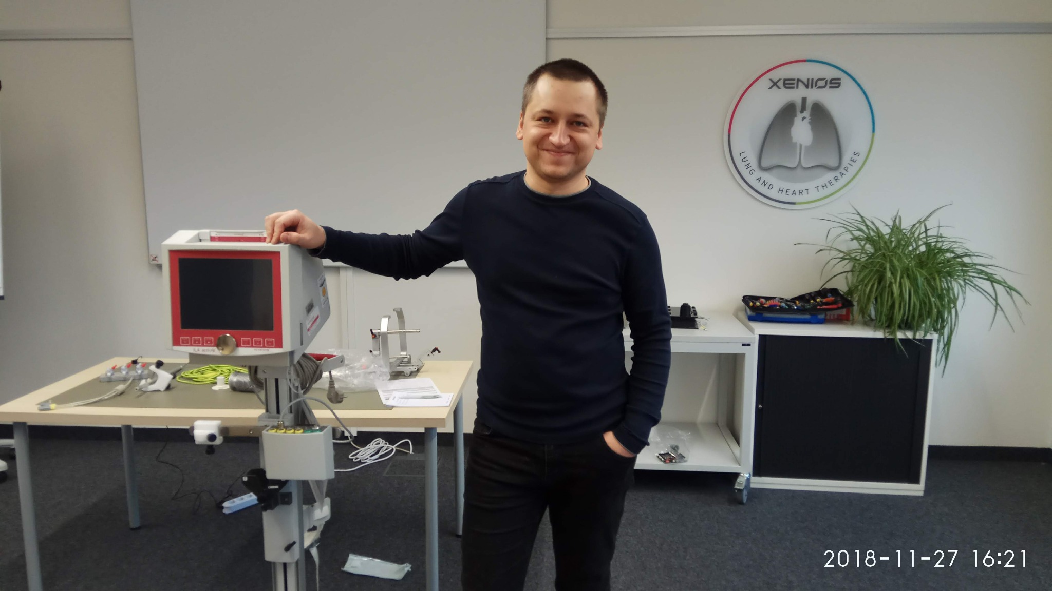 Elektronik prezentuje sprzęt medyczny Xenios: urządzenie do terapii płuc i serca na stole, z logiem firmy w tle. Specjalista z uśmiechem, gotowy do naprawy.