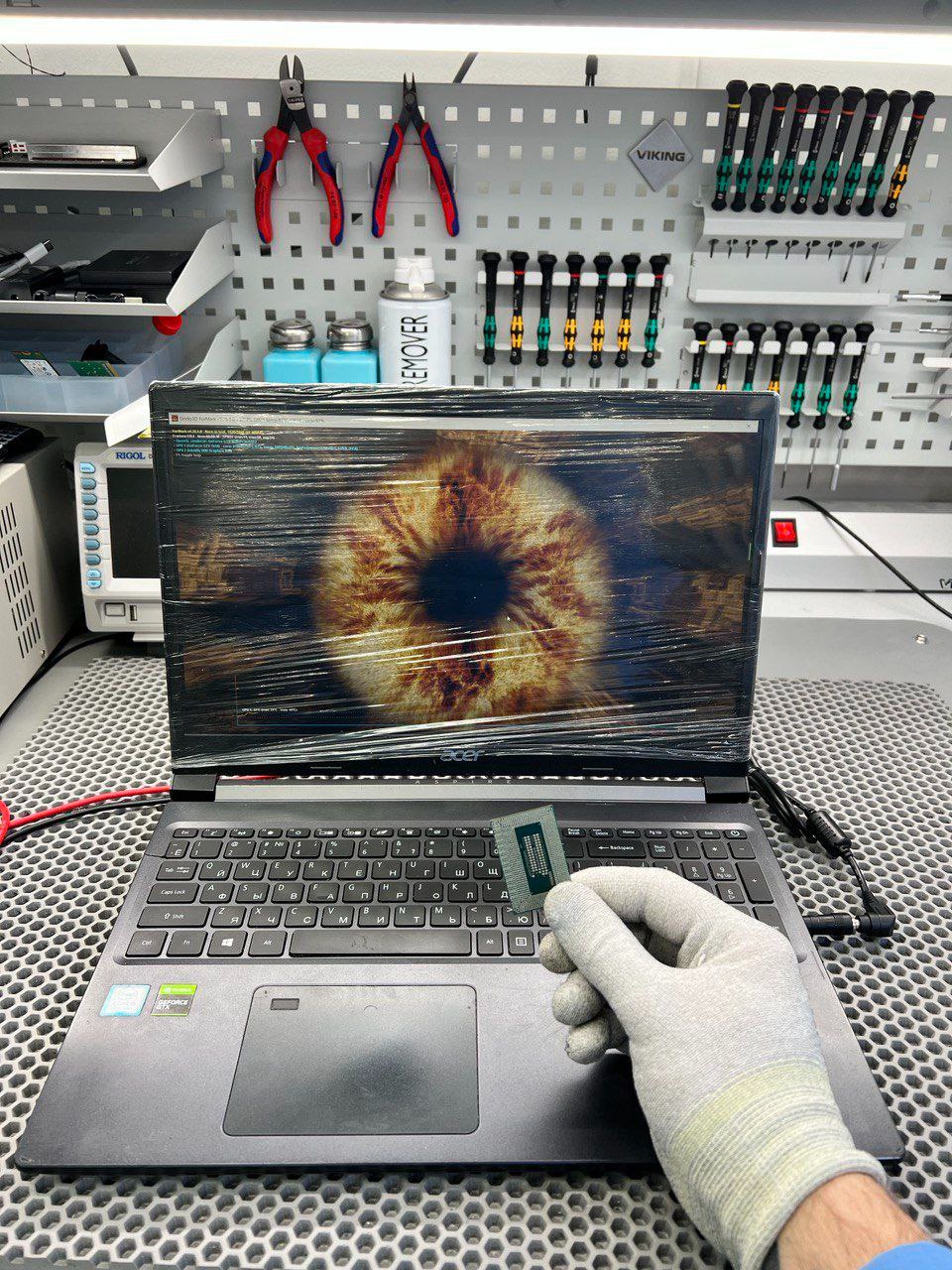 Laptop Acer, ekran z motywem oka zabezpieczony folią, dłoń w rękawicy trzyma procesor. Warsztat z narzędziami w tle. Naprawa elektroniki.