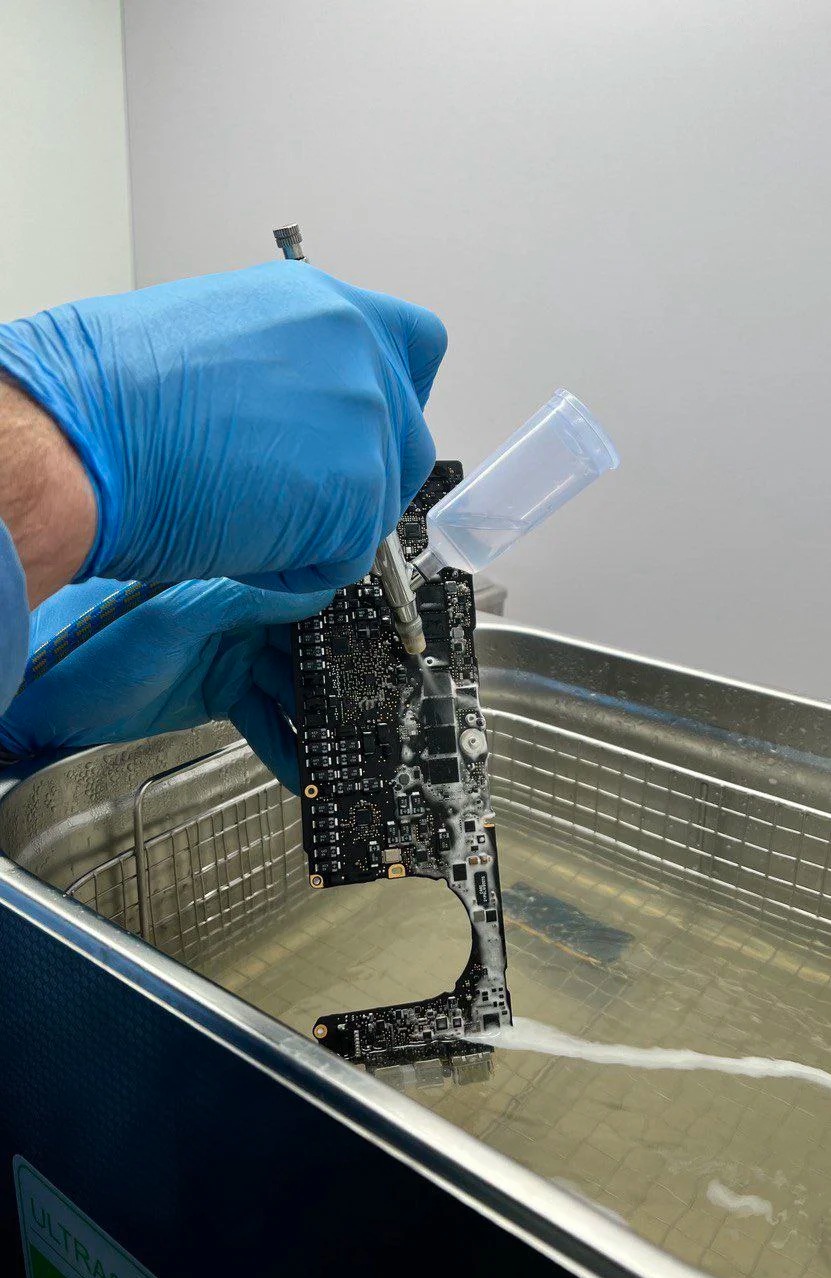 Czyszczenie płyty głównej komputera za pomocą specjalistycznego sprzętu i płynu w metalowej wannie. Płyn spływa z płyty tworząc białą smugę.