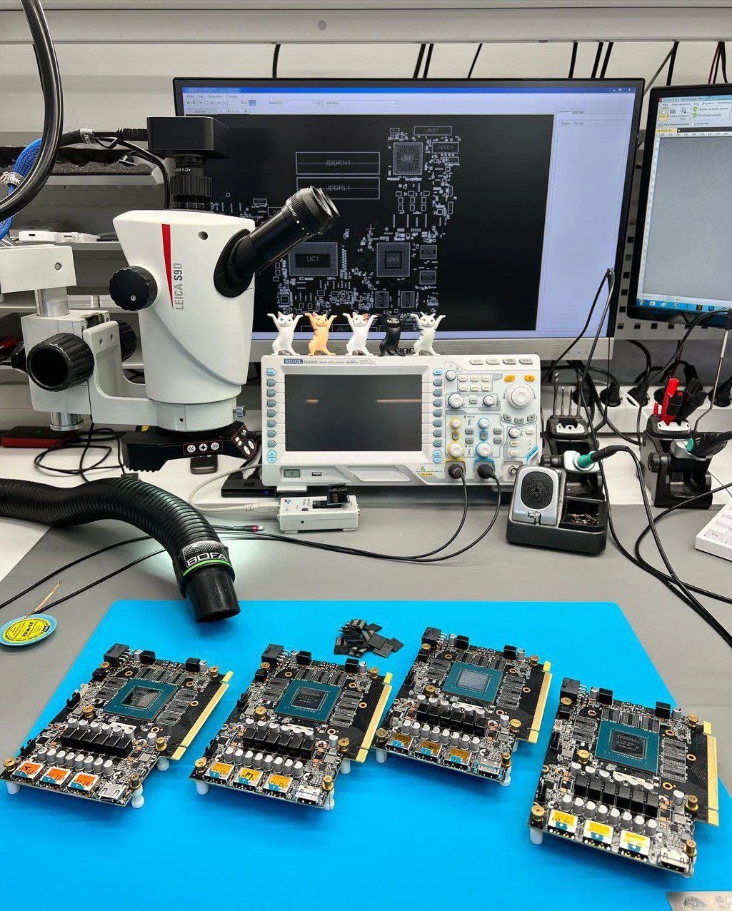 Stół serwisowy z mikroskopem, oscyloskopem i czterema kartami graficznymi na niebieskiej macie. Na monitorze widoczny schemat. Figurki kotów na oscyloskopie.