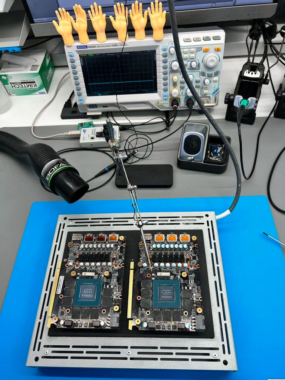 Dwie karty graficzne w ramce, podłączone do sprzętu pomiarowego, z gumowymi rękami na oscyloskopie. Sprzęt do naprawy elektroniki.