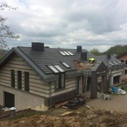 NOWAK SYSTEM Piotr Nowak - Nowoczesny dom jednorodzinny z ciemnoszarą dachówką ceramiczną, widoczne okna dachowe i kominy. Na dachu, przy krawędzi, pracuje dekarz w żółtej kamizelce. Część dachu w trakcie układania. Widoczna...