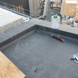 NOWAK SYSTEM Piotr Nowak - Hydroizolacja dachu płaskiego z detalami wykończeniowymi, widoczny pojemnik z klejem, szczotka i wałek, w tle pracownik i materiały budowlane.