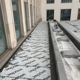 Montaż metalowych paneli dachowych na budynku, zabezpieczonych membraną z nadrukiem, widok z góry, w tle budynek z oknami.