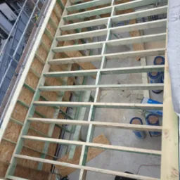 Konstrukcja drewniana dachu w trakcie budowy, widok z góry na szkielet z belek i desek, fragment rusztowania po lewej stronie, rolki izolacji termicznej widoczne w przestrzeniach między belkami...