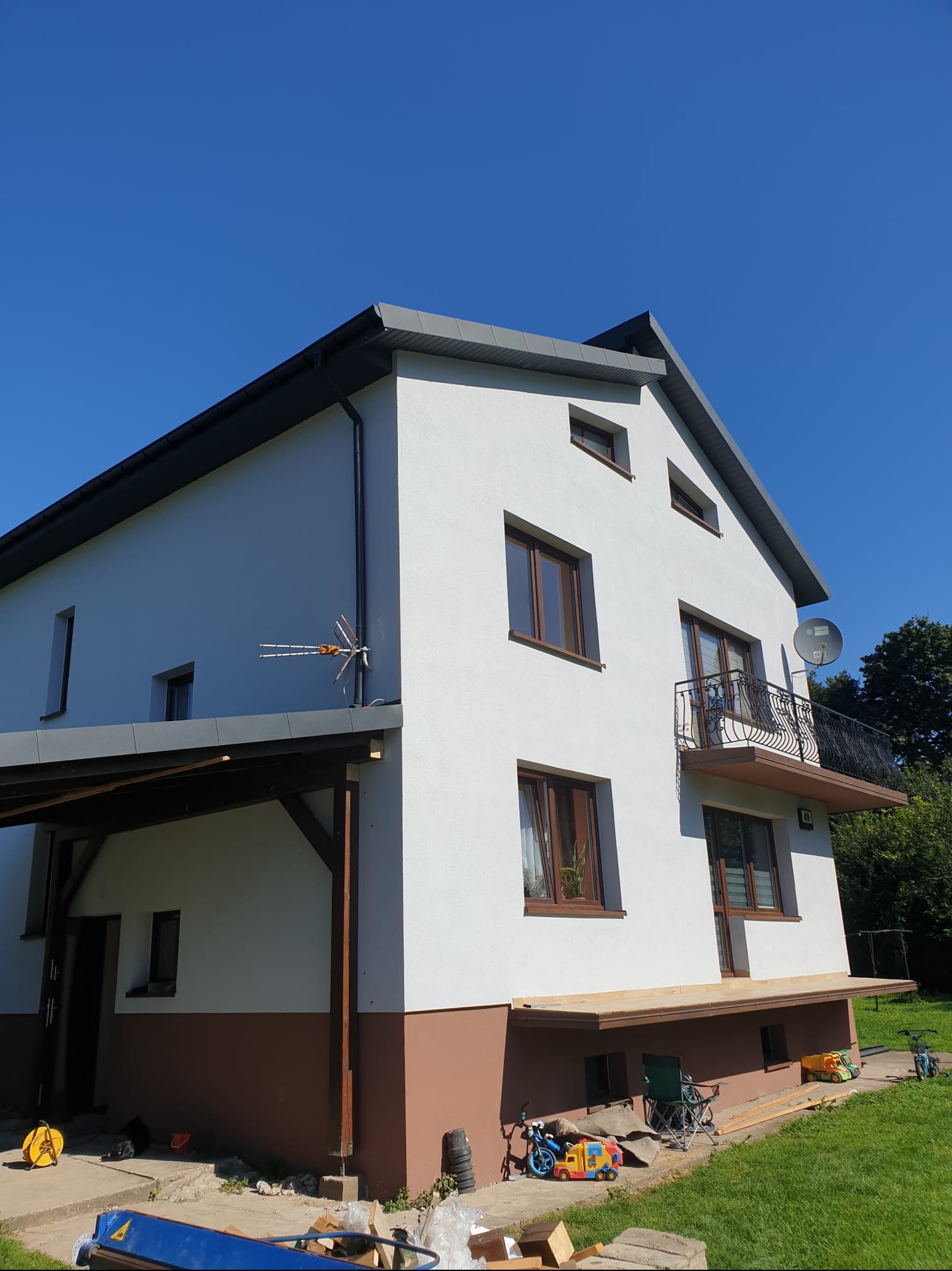 Dwupiętrowy dom z białymi ścianami i szarym dachem widziany pod kątem, z widoczną anteną na ścianie, balkonem z kutą balustradą i drewnianym tarasem na poziomie gruntu, otoczony zielenią...