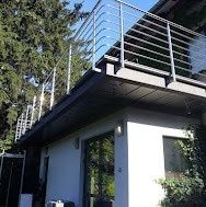 Rozbudowa domu z nowoczesną balustradą na dachu, widok z dołu na jasną elewację i zielone otoczenie.