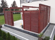 Drewniana konstrukcja z żaluzjami, prawdopodobnie osłona na śmietniki lub strefa prywatności, wykonana w Klimontowie. Widoczna kostka brukowa i elementy zieleni.