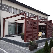 Nowoczesna pergola z ciemnego drewna przylegająca do jasnego domu z dużym oknem, widoczne donice z roślinami i fragment tarasu z szarej kostki brukowej.