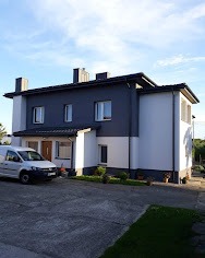 Dwukondygnacyjny dom z ciemnoszarą górną częścią elewacji i białym dołem, widoczne nowe rynny w kolorze dachu, zaparkowany biały samochód dostawczy przed budynkiem.