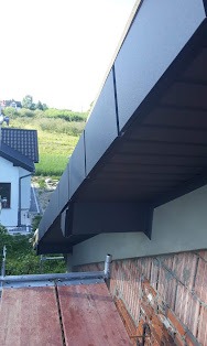 Nowoczesne, grafitowe orynnowanie zamontowane na wykończonym okapie dachu z widoczną podbitką, na tle zielonego wzgórza i zabudowań.