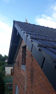 Fragment dachu z ciemną dachówką ceramiczną i czarnymi rynnami, widoczny mur z czerwonej cegły, okno bez ramy i zielone otoczenie w tle.