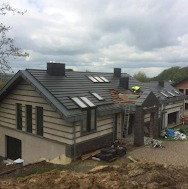 Nowoczesny dom jednorodzinny z ciemnoszarą dachówką ceramiczną, widoczne okna dachowe i kominy. Na dachu, przy krawędzi, pracuje dekarz w żółtej kamizelce. Część dachu w trakcie układania. Widoczna...