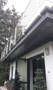 Nowoczesny balkon z metalową balustradą, widok z dołu na elewację budynku z otwartymi drzwiami, oświetlenie zewnętrzne i zieleń w tle.