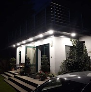 Oświetlony, parterowy dom z płaskim dachem i schodami prowadzącymi do wejścia, sfotografowany nocą. Widoczny fragment samochodu na pierwszym planie i roślinność w ogrodzie.