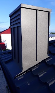 Szczegółowe ujęcie komina wentylacyjnego z czarnej blachy, zamontowanego na dachu pokrytym ciemną dachówką modułową, widok z góry, na tle zieleni i fragmentu czerwonego dachu sąsiedniego budynku.