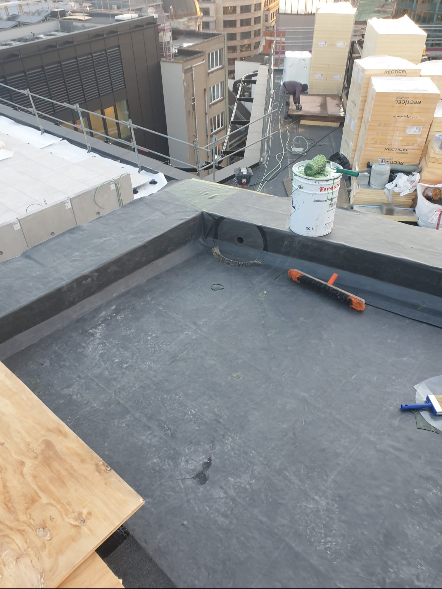 Hydroizolacja dachu płaskiego z detalami wykończeniowymi, widoczny pojemnik z klejem, szczotka i wałek, w tle pracownik i materiały budowlane.