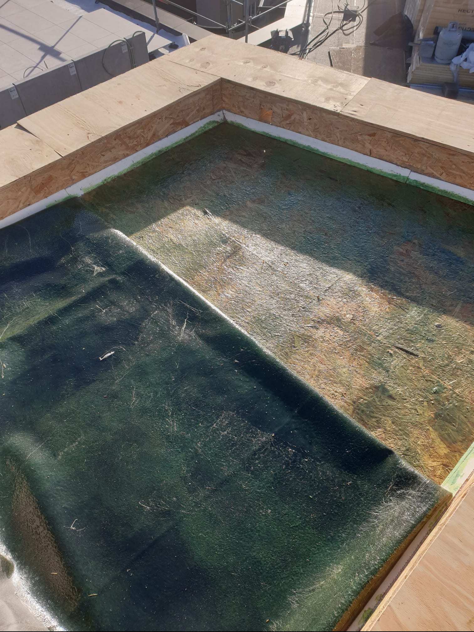 Fragment dachu w trakcie prac: widoczna płyta OSB, hydroizolacja w kolorze ciemnej zieleni, częściowo rozłożona, z widocznymi zarysowaniami i włóknami, oraz drewniana rama konstrukcyjna. W tle...