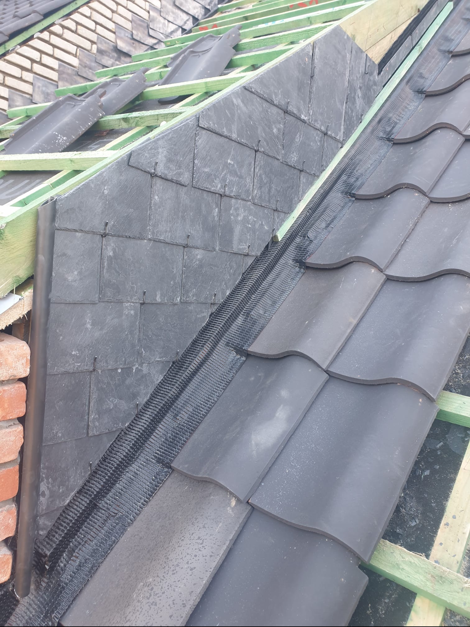 Fragment dachu w trakcie prac: połączenie łupka i dachówki ceramicznej, widoczne drewniane elementy konstrukcyjne, czarna siatka zabezpieczająca, fragment komina z cegły.