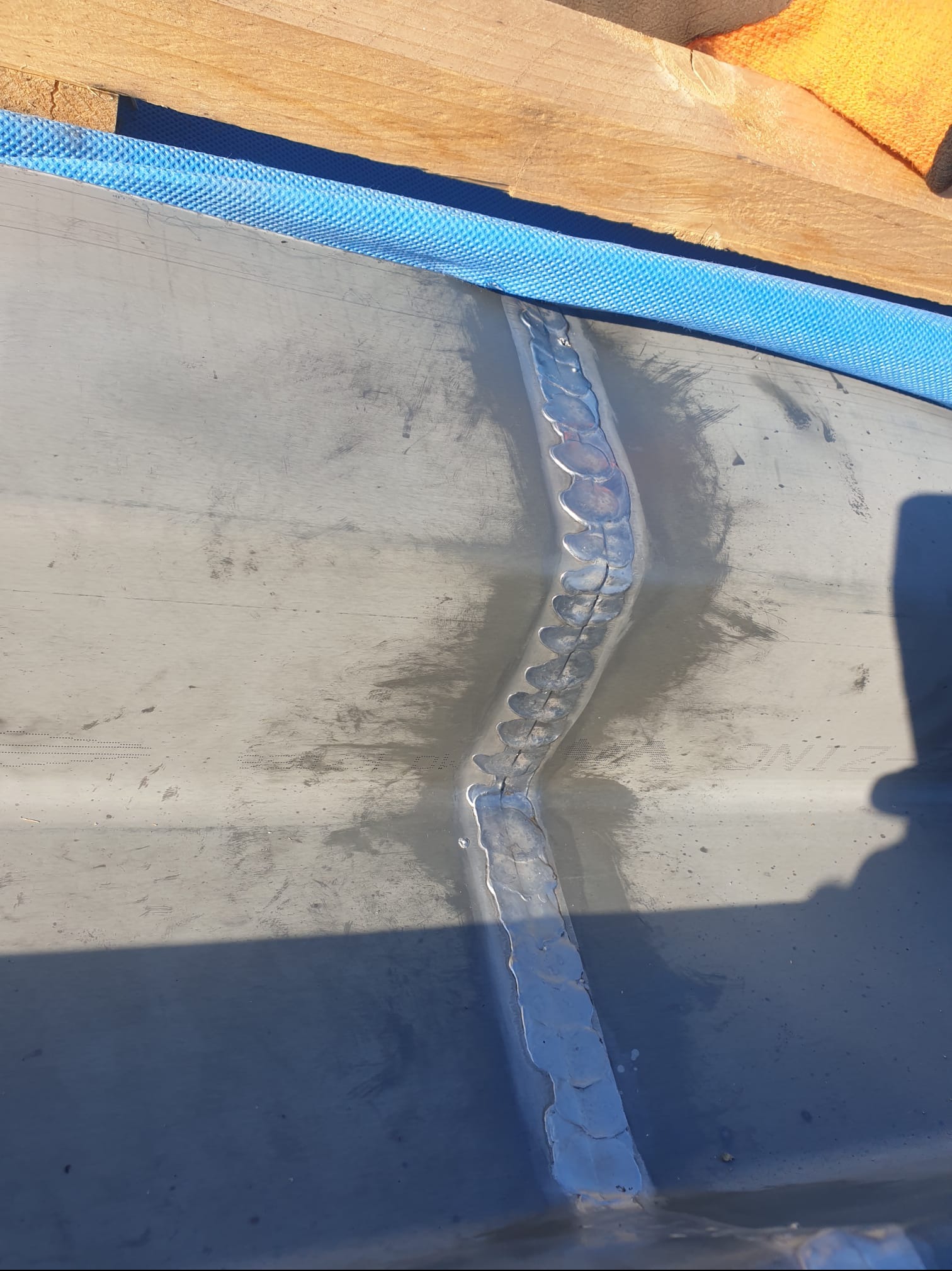 Szary metalowy dach z widocznym spawem, zabezpieczony niebieską membraną wzdłuż krawędzi i fragmentem drewnianej konstrukcji.