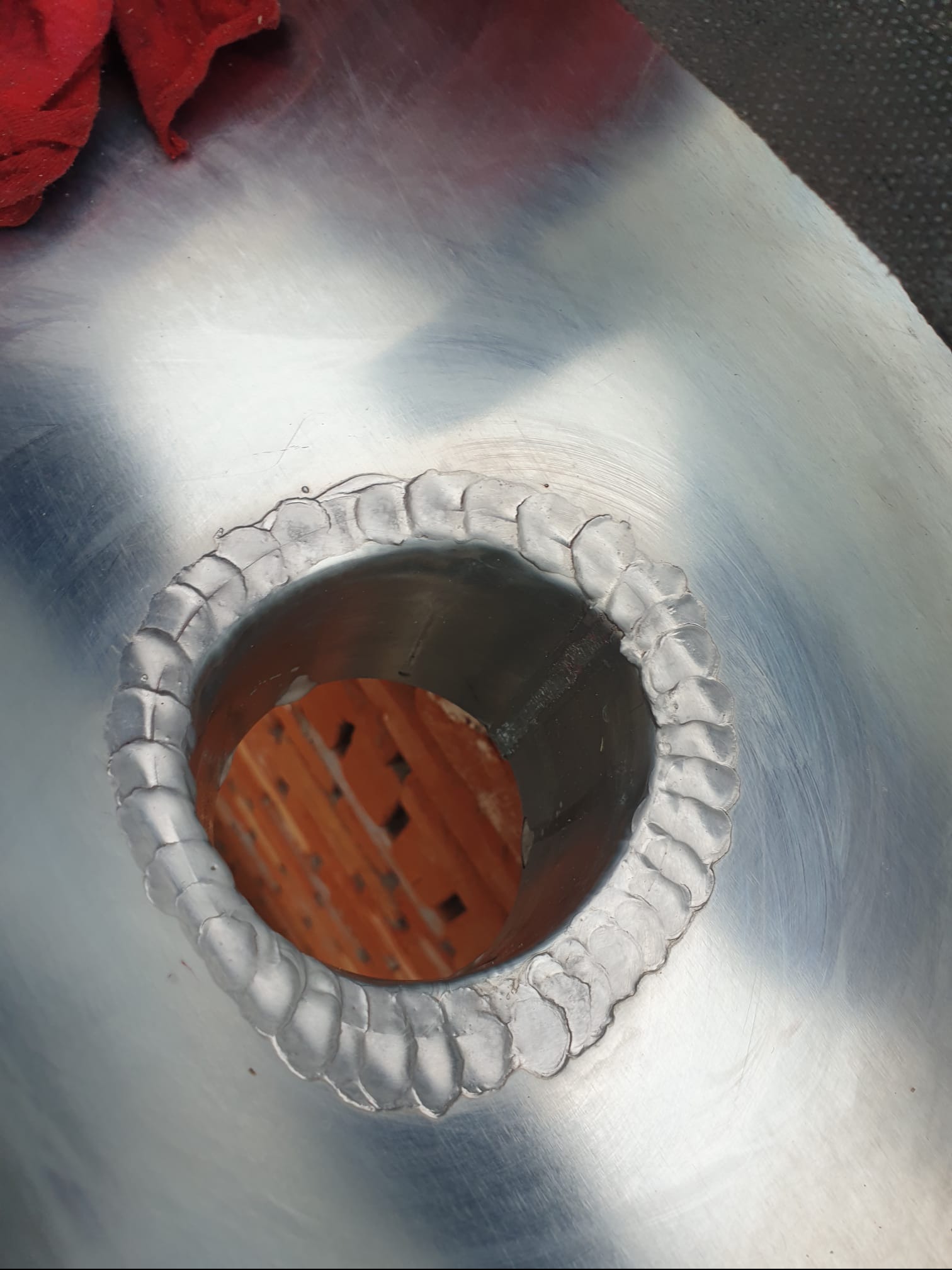 Szczegółowe ujęcie spawu wokół okrągłego otworu w metalowej powierzchni, z widoczną cegłą wewnątrz otworu i śladami obróbki na metalu.