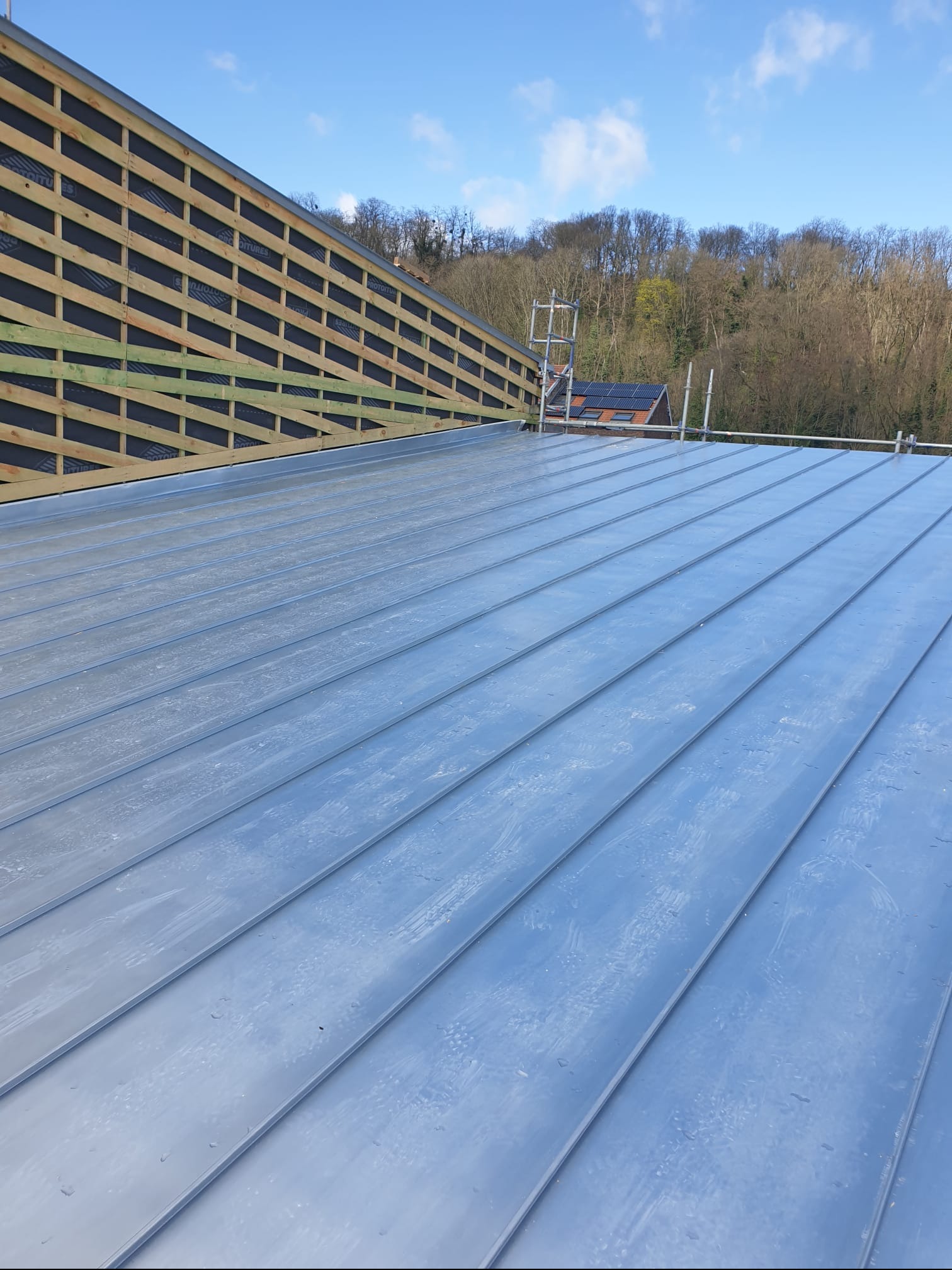 Nowe, metalowe pokrycie dachu z widoczną konstrukcją drewnianą z membraną oraz rusztowaniem w tle, w słoneczny dzień.