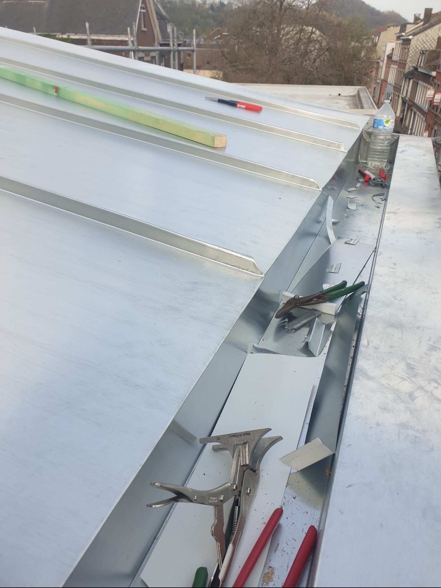 Metalowy dach w trakcie montażu rynny, widoczne narzędzia dekarskie, poziomica, butelka wody i elementy blacharskie.