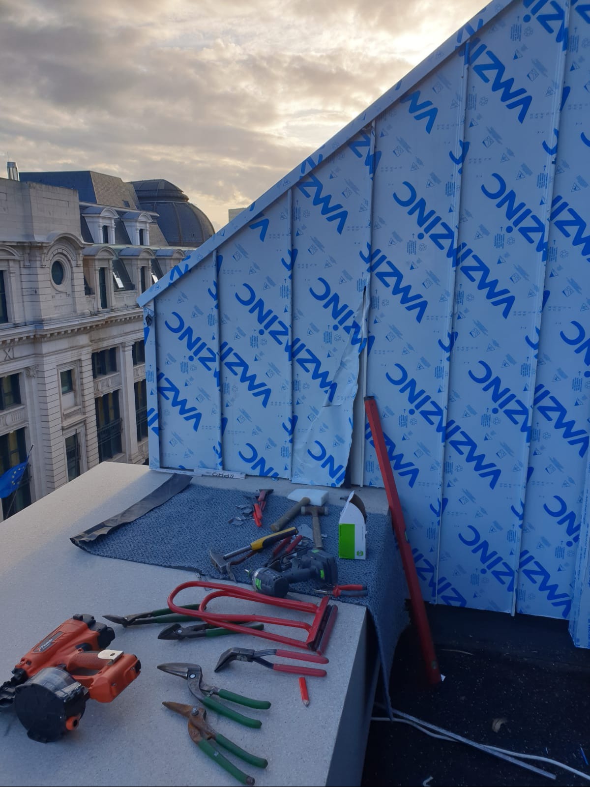 Montaż paneli z blachy tytan-cynk na dachu skośnym, widoczne narzędzia dekarskie i częściowo usunięta folia ochronna z logo producenta, w tle budynek miejski.