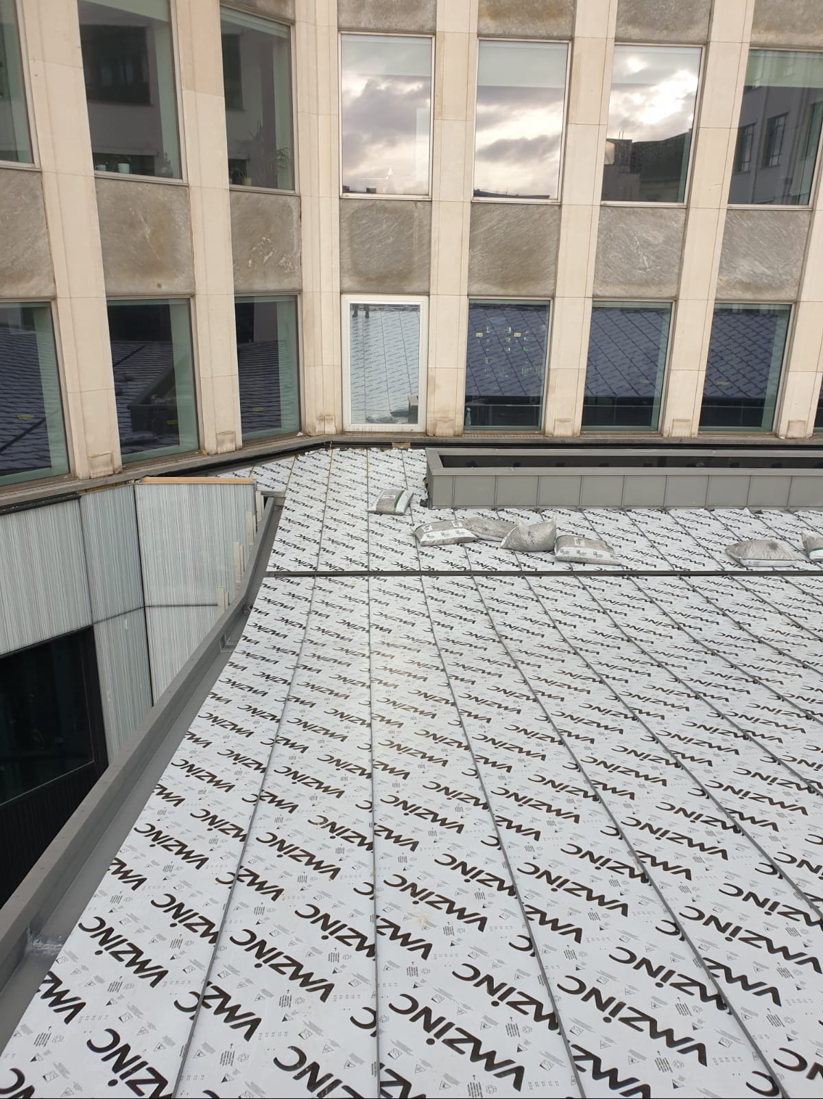 Nowe pokrycie dachu z blachy trapezowej z nadrukiem producenta, widok z góry na dach budynku biurowego z odbiciami w oknach.