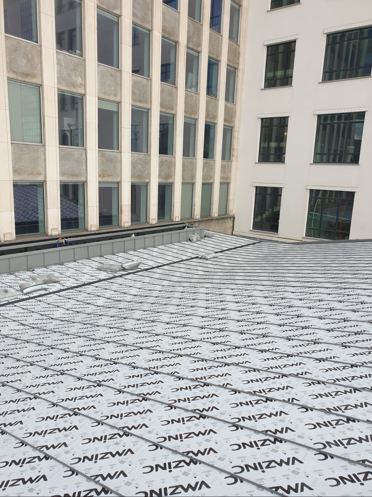 Nowa warstwa izolacyjna z nadrukiem na dachu budynku wielorodzinnego, widok z góry na przygotowany dach przed kryciem, w tle budynki biurowe.