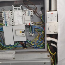 TK&ELECTRICS - Otwarta skrzynka elektryczna z bezpiecznikami, przewodami i oznaczeniem ostrzegawczym 'Danger 400 Volts' na szarej obudowie, obok białe gniazdka i rurki instalacyjne.