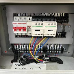 TK&ELECTRICS - Otwarta skrzynka elektryczna z bezpiecznikami, przekaźnikami i kolorowymi przewodami. Widoczne oznaczenia L1, L2, L3, N na kartce. Instalacja w szarej obudowie.