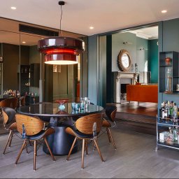 TK&ELECTRICS - Stylowe wnętrze jadalni z okrągłym stołem, krzesłami z drewna i czarnej skóry, designerską lampą i barkiem na kółkach, widok na salon z kominkiem i pomarańczową sofą.