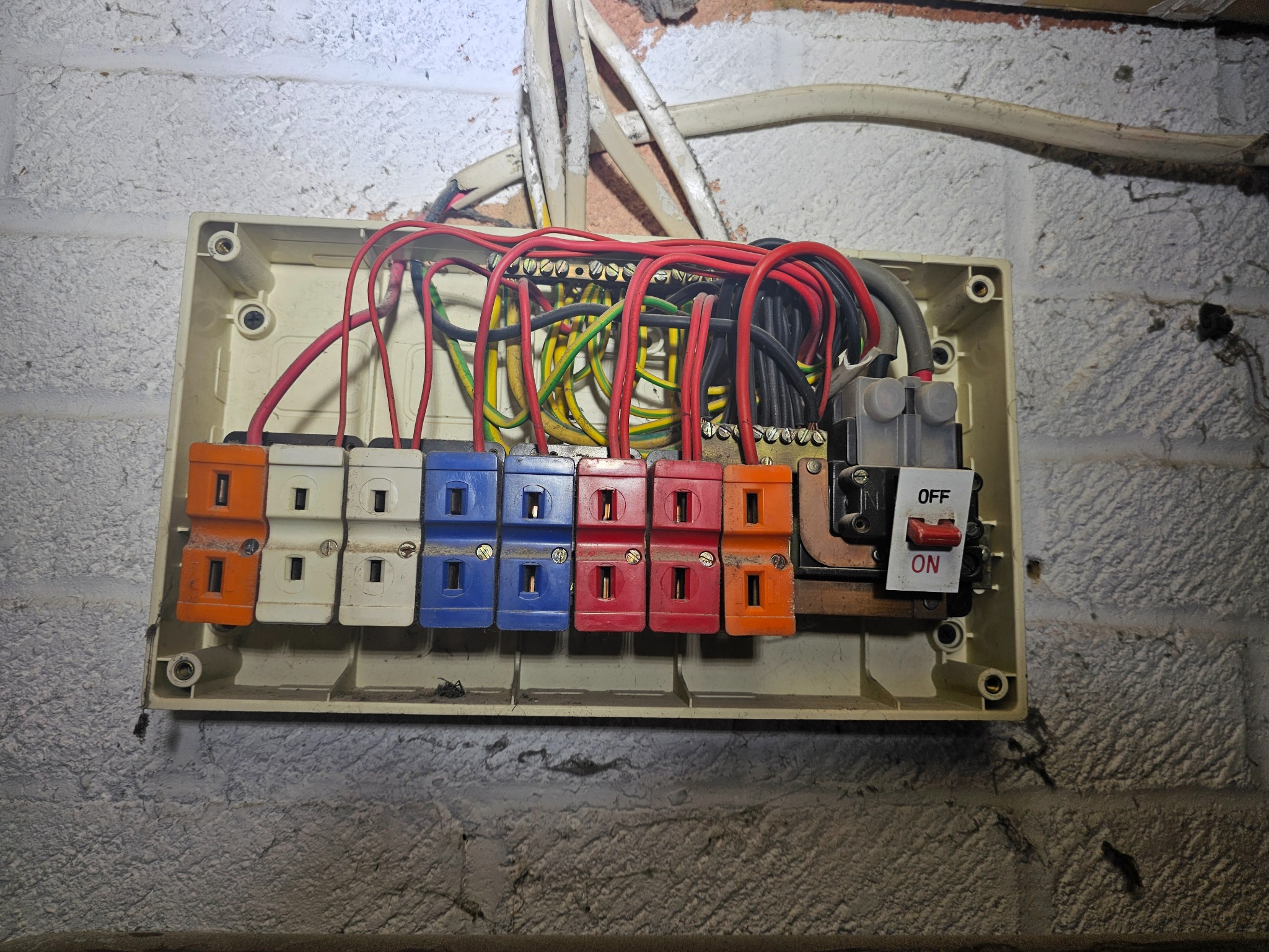 Otwarta skrzynka bezpiecznikowa z kolorowymi wtyczkami i przewodami na szorstkiej ścianie z cegieł. Widoczny przełącznik w pozycji 'ON'.