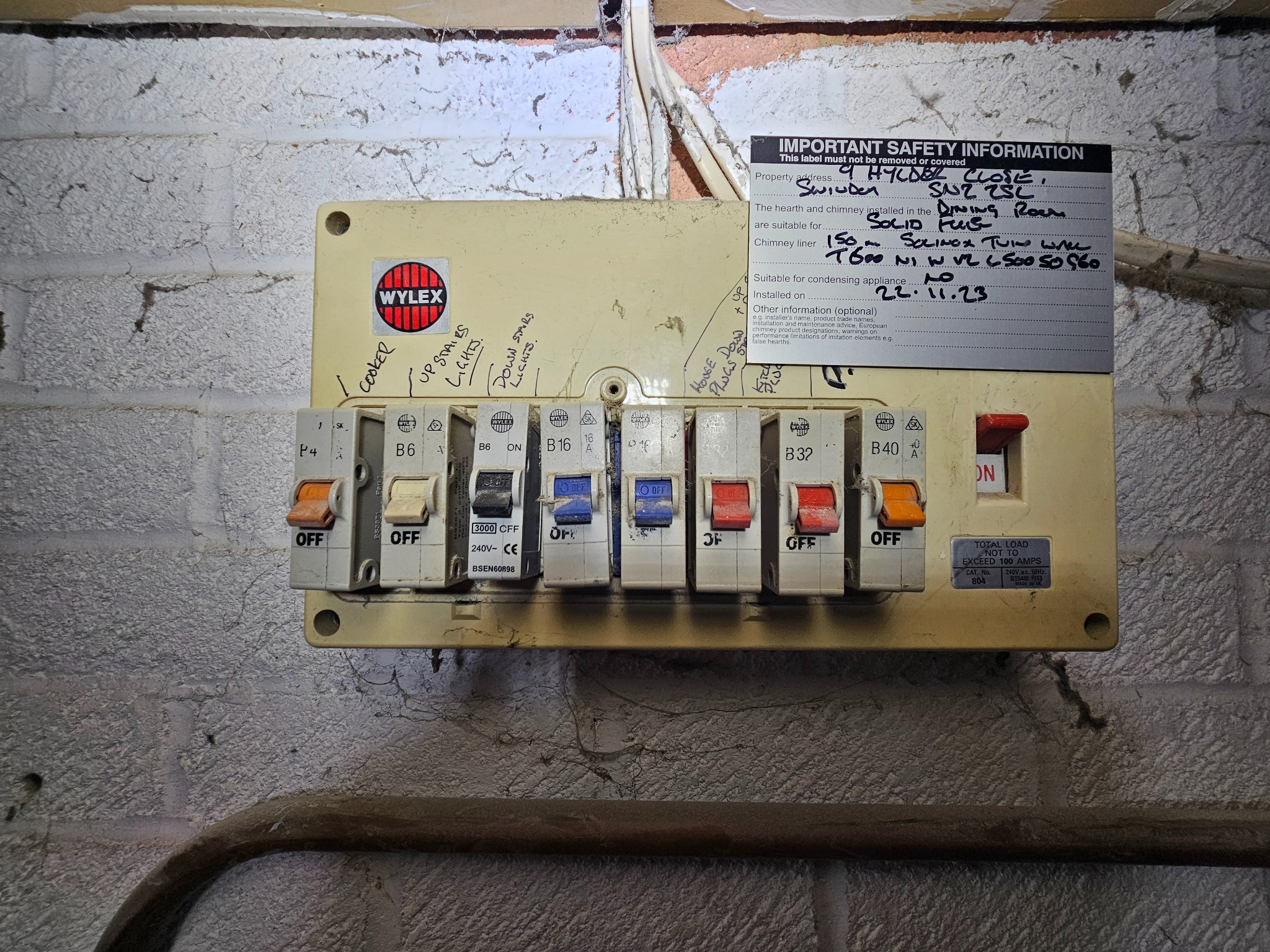 Tablica bezpiecznikowa Wylex na ścianie z cegły, z widocznymi przełącznikami i naklejką z informacjami o instalacji elektrycznej. Widoczne ślady użytkowania.