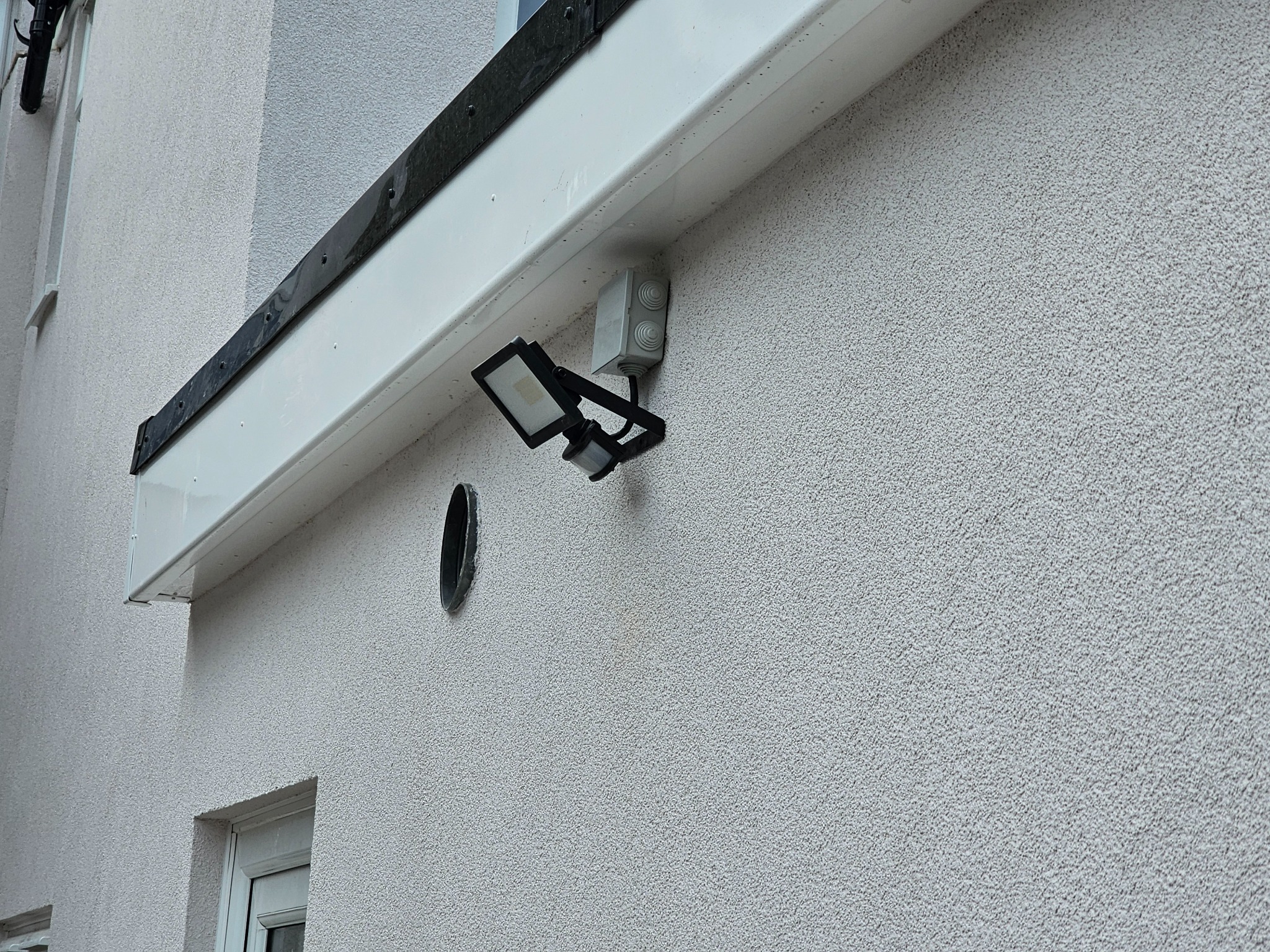 Zamontowany zewnętrzny reflektor LED z czujnikiem ruchu na elewacji budynku o szarej, ziarnistej strukturze. Widoczna puszka instalacyjna i fragment okna.