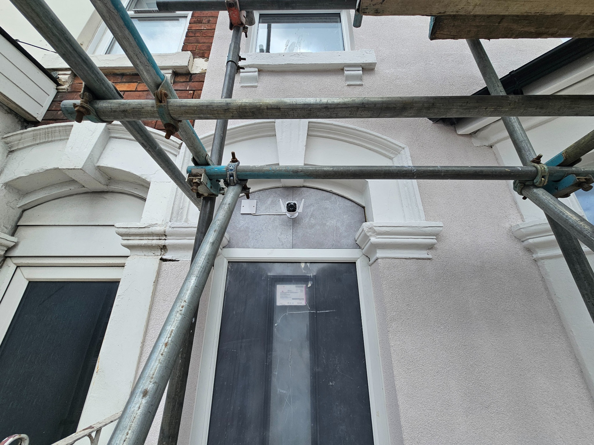 Elewacja domu w trakcie prac remontowych, widoczne rusztowanie, okno i zamontowana kamera monitoringu. Jasna fasada z dekoracyjnymi elementami architektonicznymi.