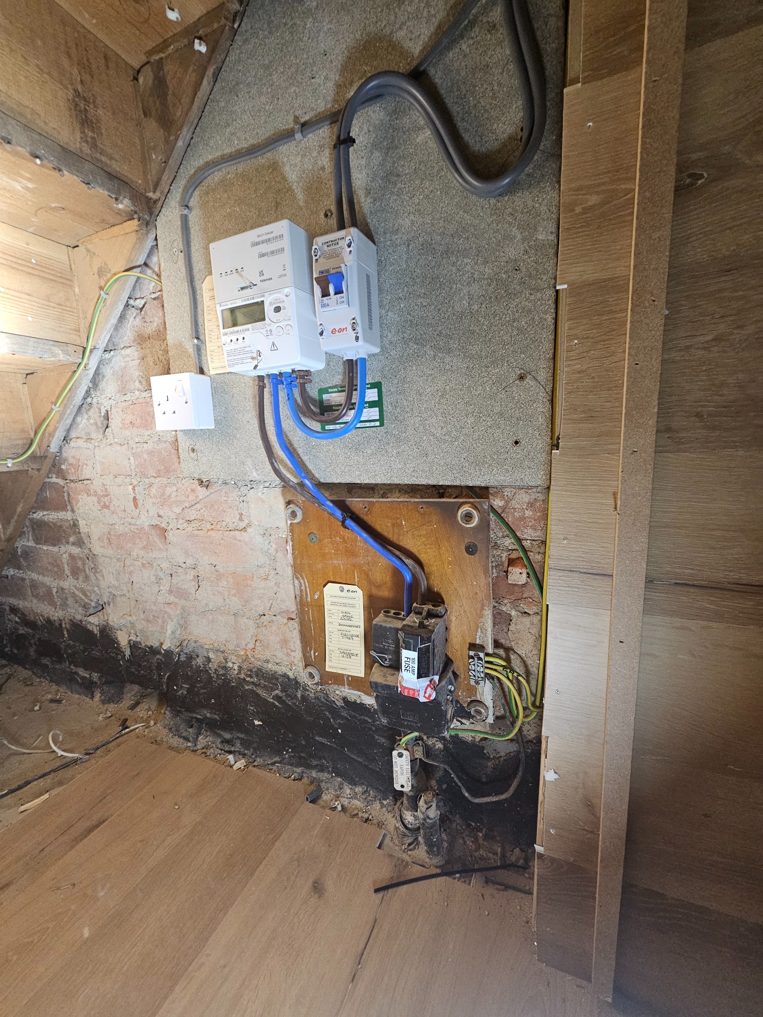 Zamontowany licznik elektryczny E-on z zabezpieczeniami w pomieszczeniu z surową cegłą i drewnianą podłogą. Widoczne kable i gniazdko.