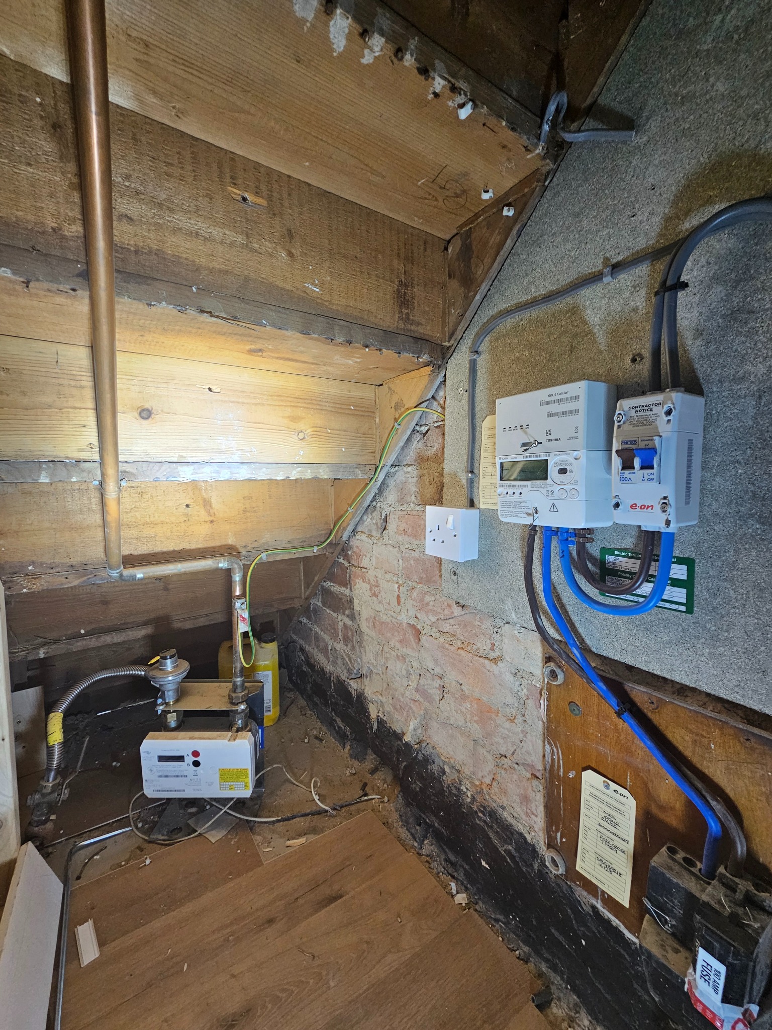 Nowy licznik elektryczny i gazowy zamontowany w rogu pomieszczenia z odsłoniętą cegłą i drewnianą konstrukcją. Widoczne przewody i rury.