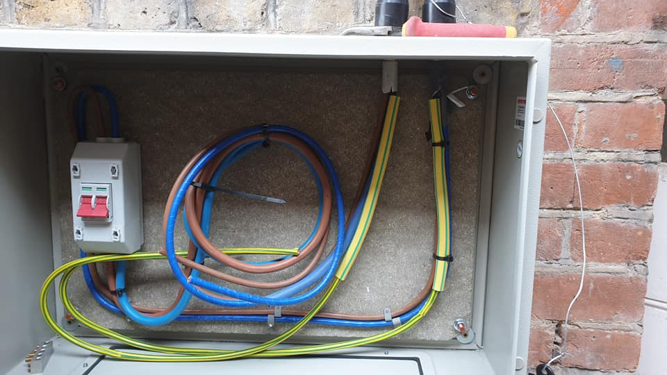 Rozdzielnica elektryczna z okablowaniem wewnątrz, widoczne przewody w kolorach niebieskim, brązowym i żółto-zielonym, zamontowany wyłącznik nadprądowy.