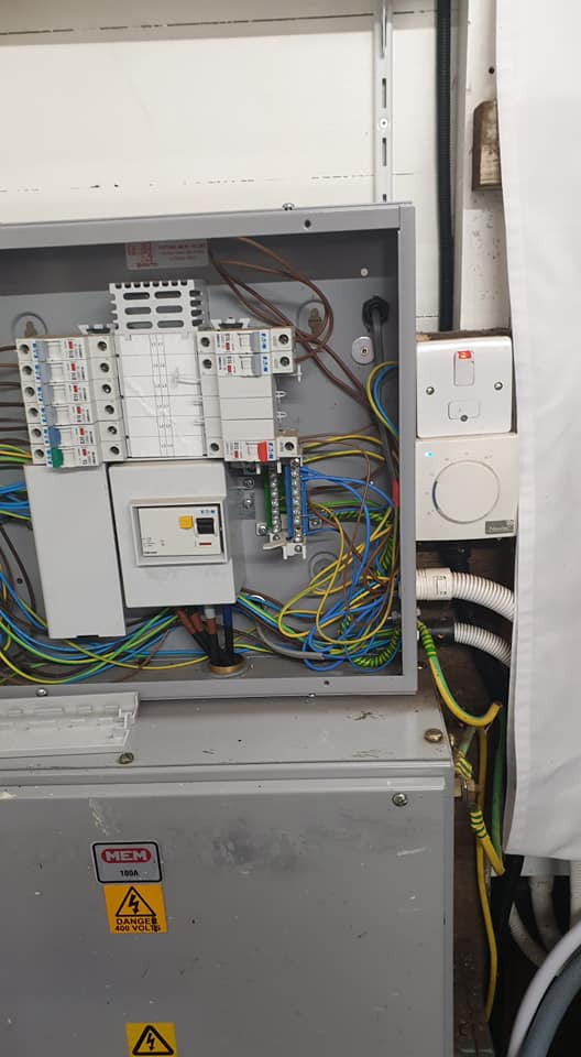 Otwarta skrzynka elektryczna z bezpiecznikami, przewodami i oznaczeniem ostrzegawczym 'Danger 400 Volts' na szarej obudowie, obok białe gniazdka i rurki instalacyjne.