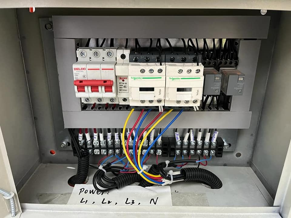 Otwarta skrzynka elektryczna z bezpiecznikami, przekaźnikami i kolorowymi przewodami. Widoczne oznaczenia L1, L2, L3, N na kartce. Instalacja w szarej obudowie.