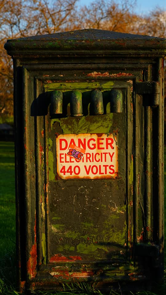 Stara, zielona skrzynka elektryczna z napisem 'Danger Electricity 440 Volts' na tle trawy i drzew. Widoczne ślady rdzy i odpryski farby.