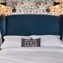 Nastrojowa sypialnia z tapetą w motywy karciane, niebieskim zagłówkiem i poduszką z napisem 'New York Paris Balham'. Dwa kinkiety oświetlają przestrzeń.