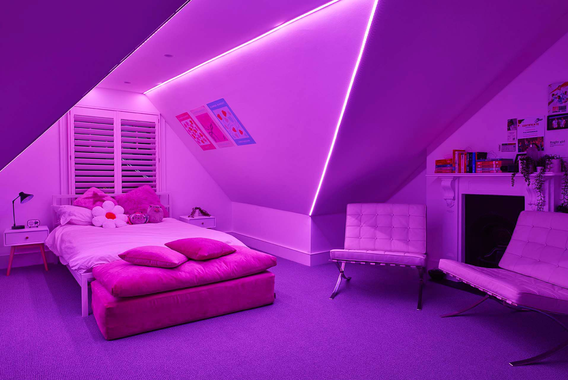 Pokój na poddaszu z oświetleniem LED w kolorze fioletowym, łóżkiem, stolikami nocnymi i fotelami. Widoczne skosy dachu i elementy dekoracyjne.