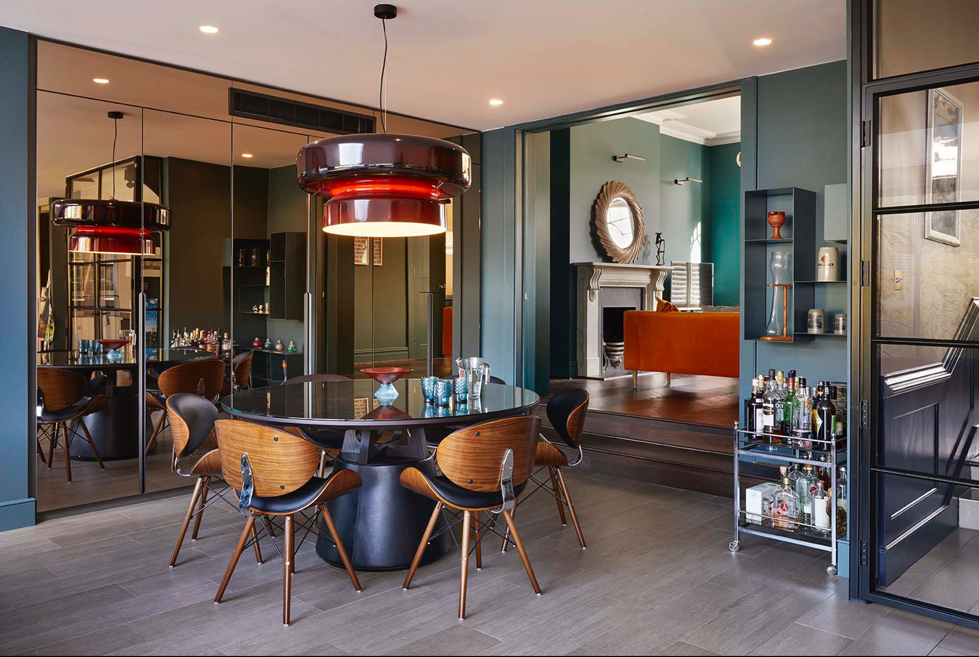 Stylowe wnętrze jadalni z okrągłym stołem, krzesłami z drewna i czarnej skóry, designerską lampą i barkiem na kółkach, widok na salon z kominkiem i pomarańczową sofą.